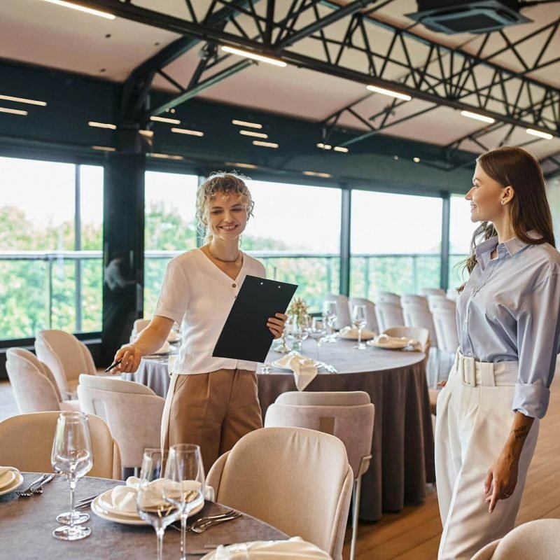 event-organizer-with-clipboard-pointing-at-table-with-festive-setting-near-woman-in-banquet-hall.jpg